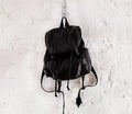 Black drawstring rucksack with long straps hanging from a hook on a white textured wall