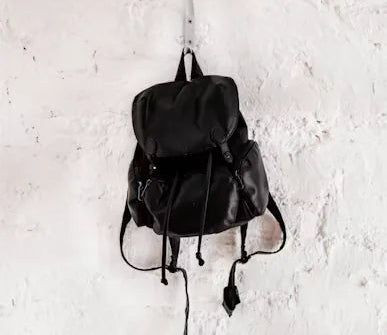 Black drawstring rucksack with long straps hanging from a hook on a white textured wall