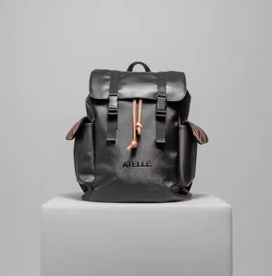 Black leather rucksack backpack with a buckled flap, tan drawcord and side pockets, on a white plinth
