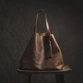 Brown leather tote bag with long handles and a tassel, on a wooden stool against a dark textured wall
